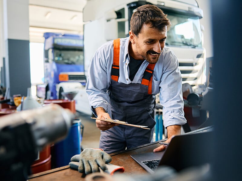 Happy mechanic using laptop while working at truck repair workshop